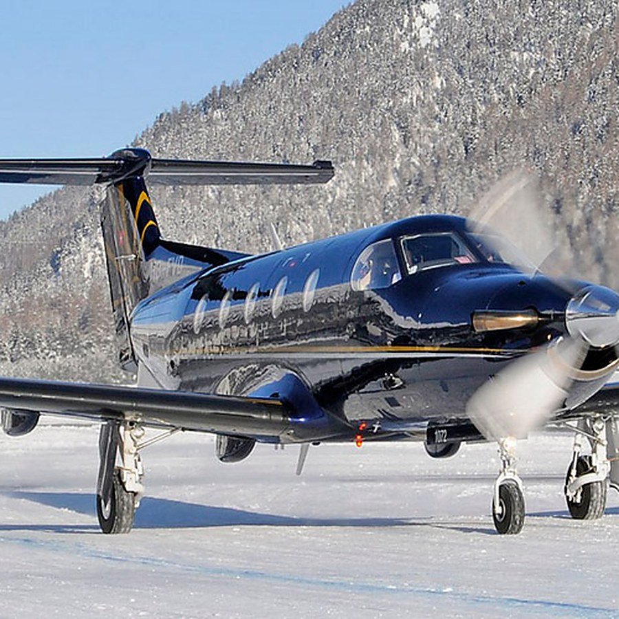 Un PC-12 en una pista de montaña nevada, con bosques y cumbres detrás.